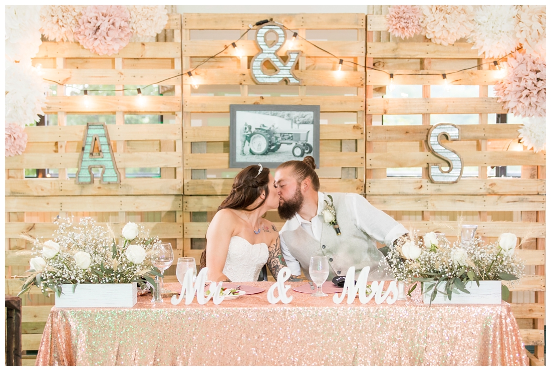 Bride and Groom kissing at sweetheart table with a pallet wall. Reception details and decor. Rustic wedding decor. Rose gold wedding decor. Maryland wedding at Circle D Farm in Woodbine. Maryland Wedding Photographer.